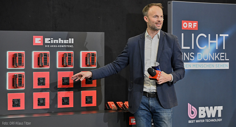 A man presents Einhell batteries on a wall display next to an ORF “Licht ins Dunkel” banner.