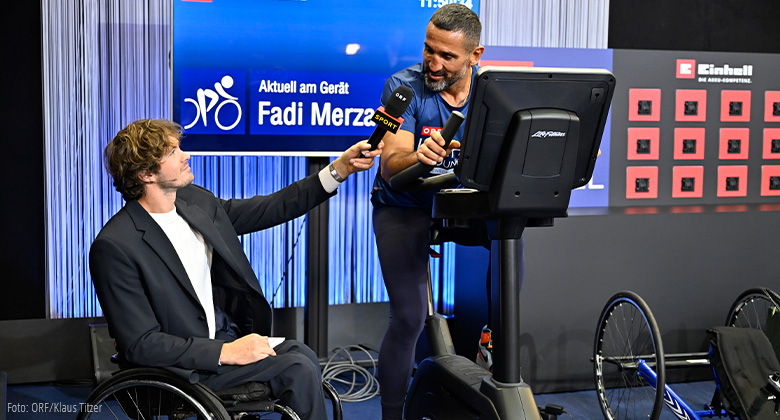 A man in a wheelchair hands a microphone to an athlete on an exercise bike; “Fadi Merza” is displayed in the background.