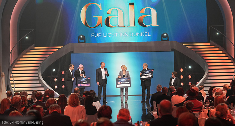 Stage scene of the ORF charity gala "Licht ins Dunkel" with five people presenting donation amounts on large boards.