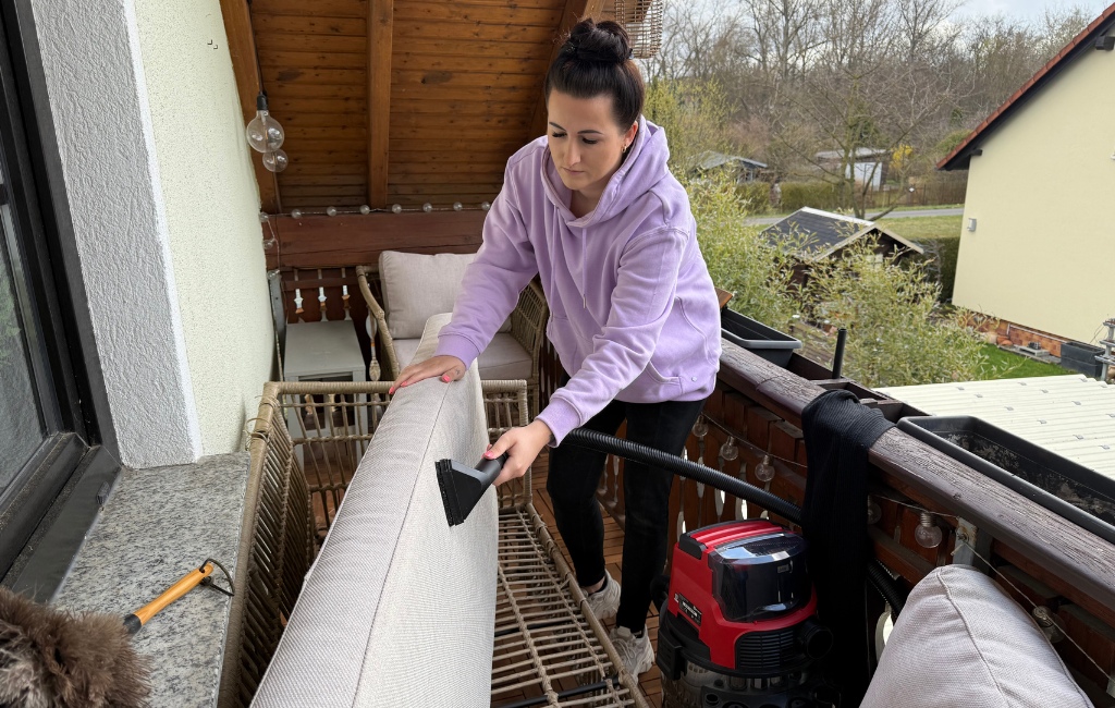 Eine Frau reinigt mit der Polsterdüse eines rot-schwarzen Einhell Nass-Trockensaugers die großen, grauen Kissen einer Balkon-Lounge.