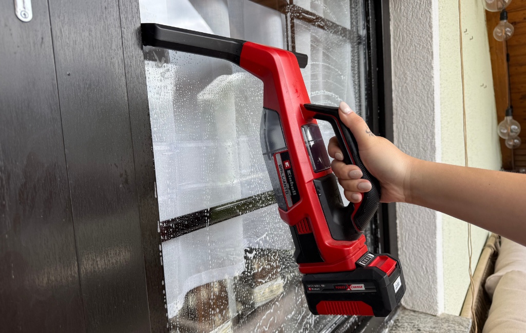 Eine Hand führt den rot-schwarzen Einhell Akku-Fensterreiniger BRILLIANTO über eine schäumende Fensterscheibe.
