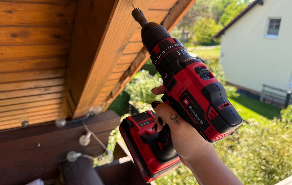 Nahaufnahme einer Hand, die mit einem rot-schwarzen Einhell Akku-Bohrschrauber eine Schraube in einen hölzernen Dachbalkon dreht.