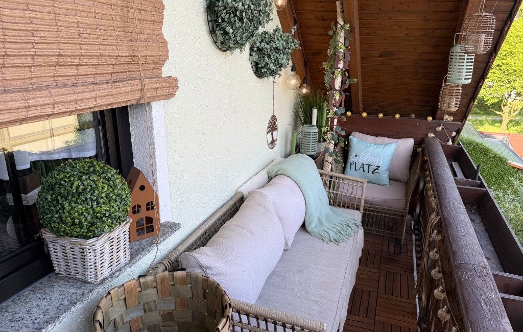 Ein fertig dekorierter, gemütlicher Balkon mit hellen Lounge-Möbeln, Holzboden, Lichterketten und Pflanzen bei Tageslicht.