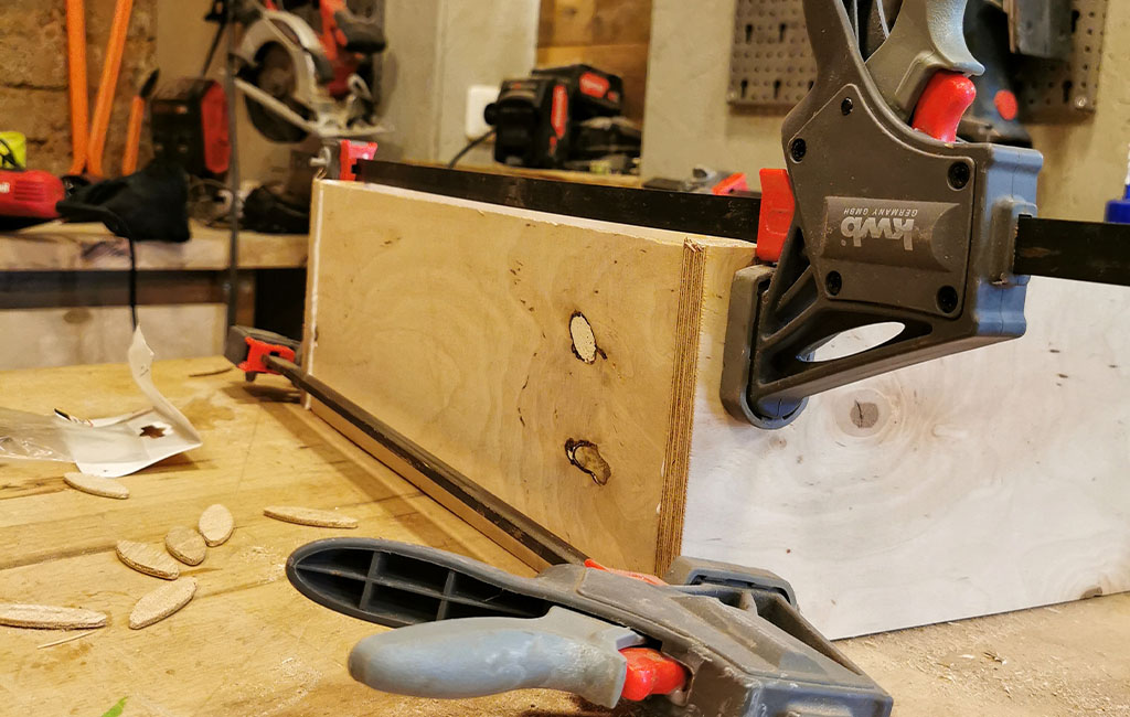 Zwei Holzplatten werden mit Schraubzwingen und Holzleim zusammengepresst. Holzverbindungsplättchen liegen daneben.