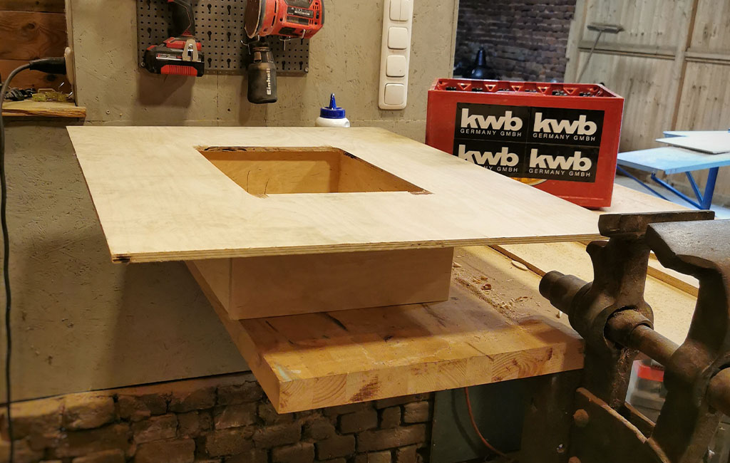 Eine Holzplatte mit viereckiger Öffnung liegt auf einem Holzkasten. Im Hintergrund eine Werkzeugwand und eine kwb-Getränkekiste.