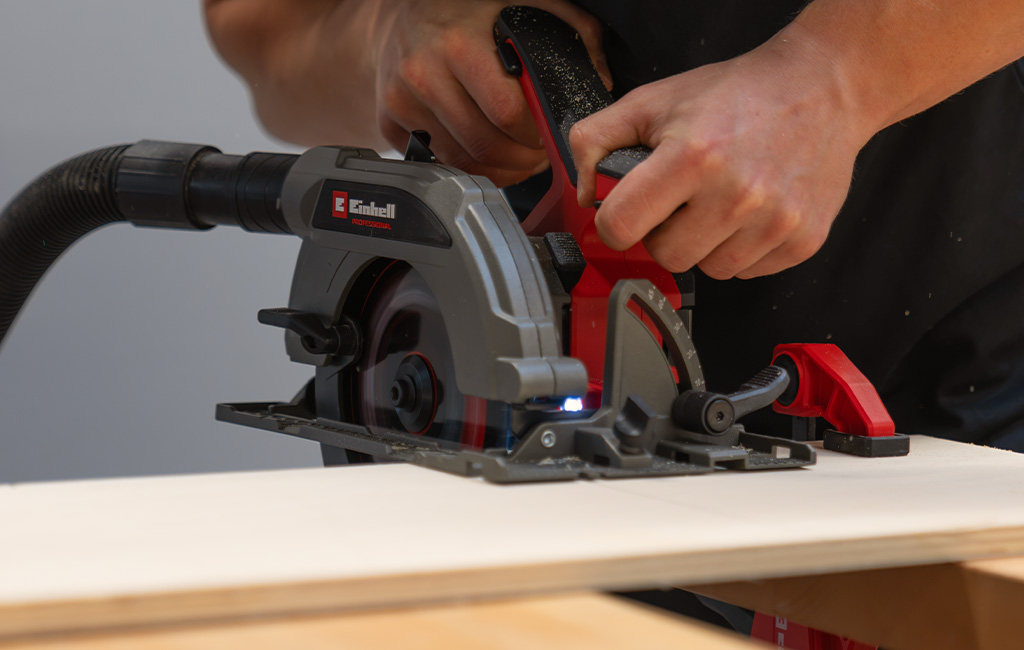 A person uses an Einhell circular saw to cut a wooden board; a dust-extraction hose is attached.