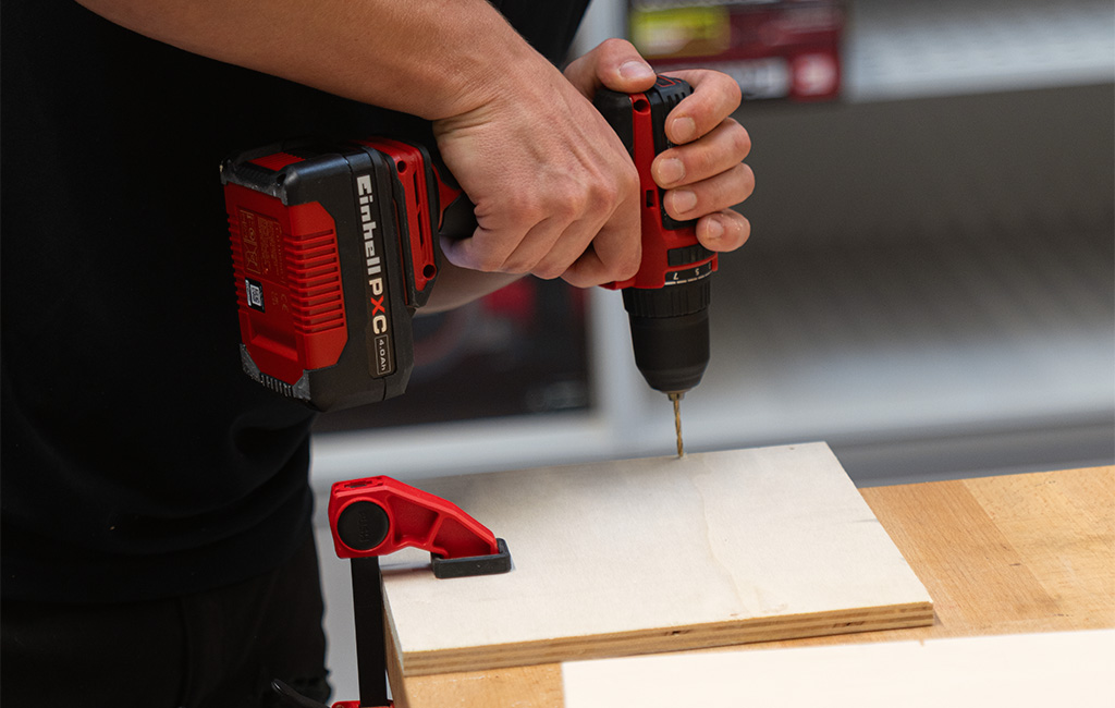 A person uses an Einhell cordless drill to drill a hole in a wooden board secured with a clamp.