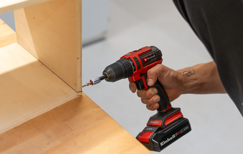 A person uses an Einhell cordless drill to drill into a wooden cabinet joint.