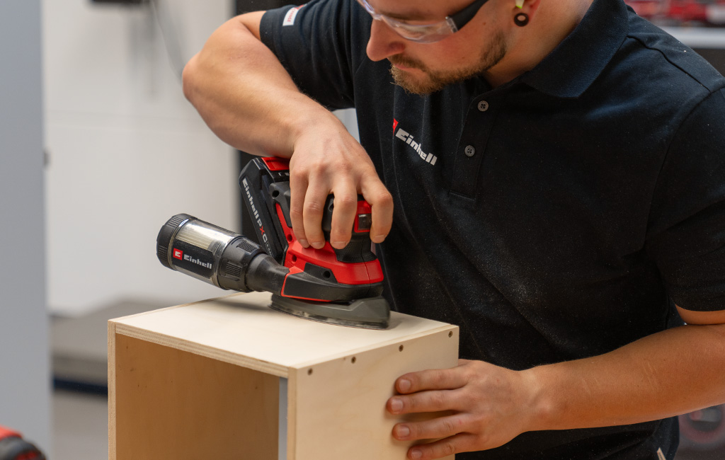 A person uses an Einhell cordless sander to sand the top of a wooden box.