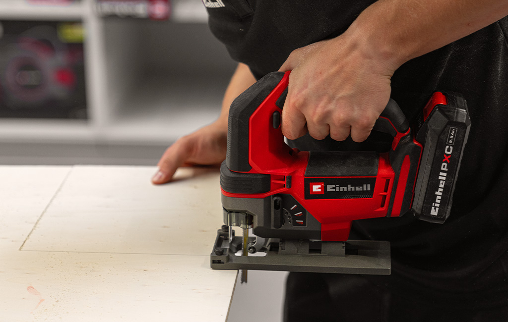 A person uses an Einhell cordless jigsaw to cut a wooden board along a marked line.