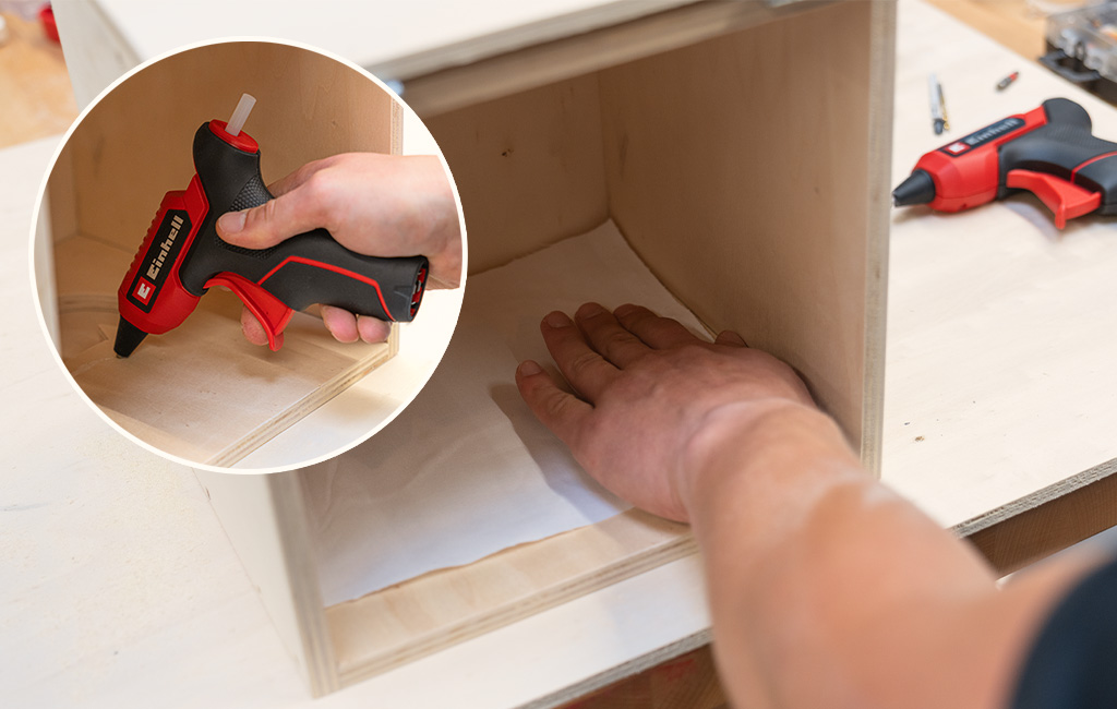 A person secures a fabric cloth inside a wooden box; the inset shows an Einhell cordless hot glue gun applying adhesive.