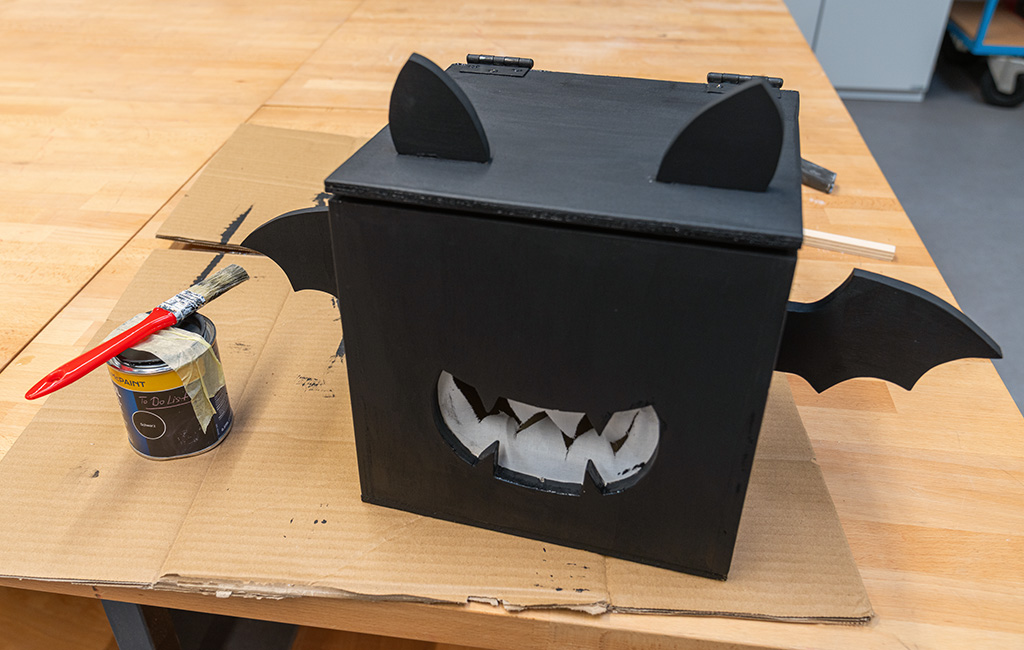 Finished black wooden bat box with ears, wings and a white jagged-mouth cutout, next to a paint can and brush.