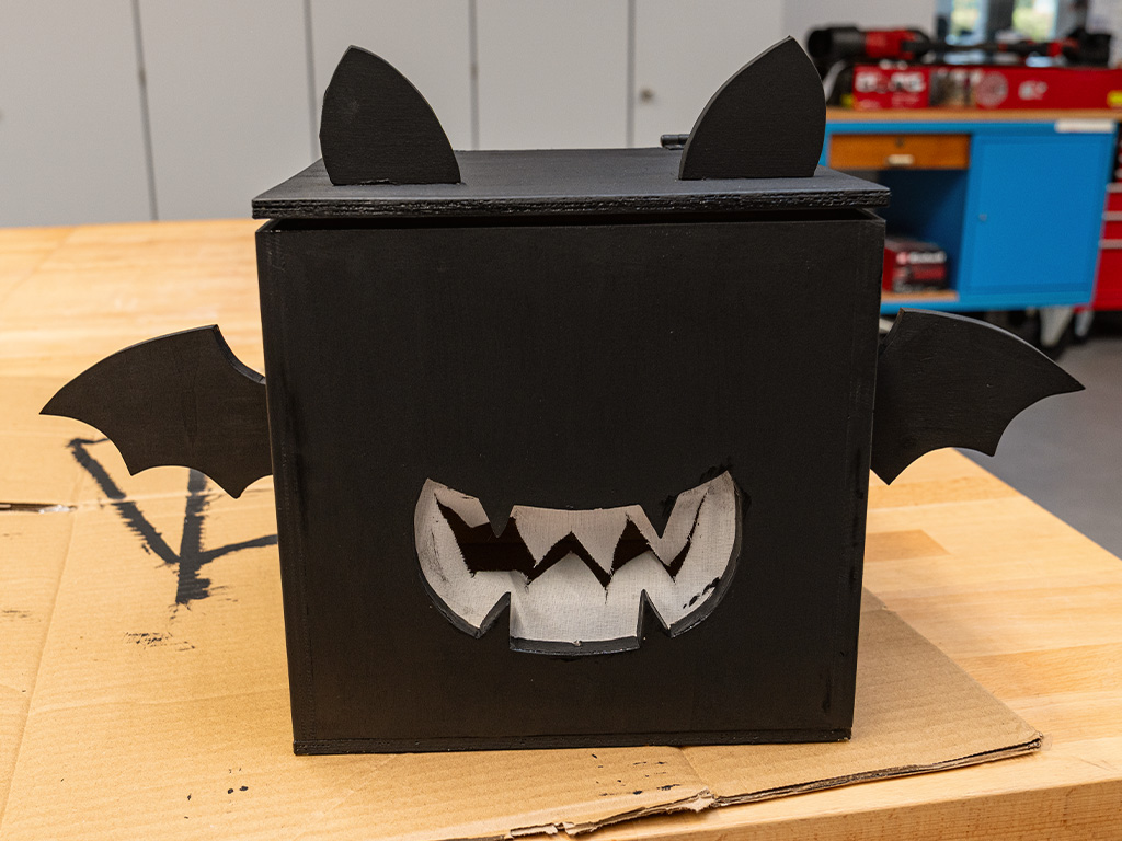 Black wooden bat box with ears, wings and a white jagged-toothed mouth, standing on cardboard on a workbench.
