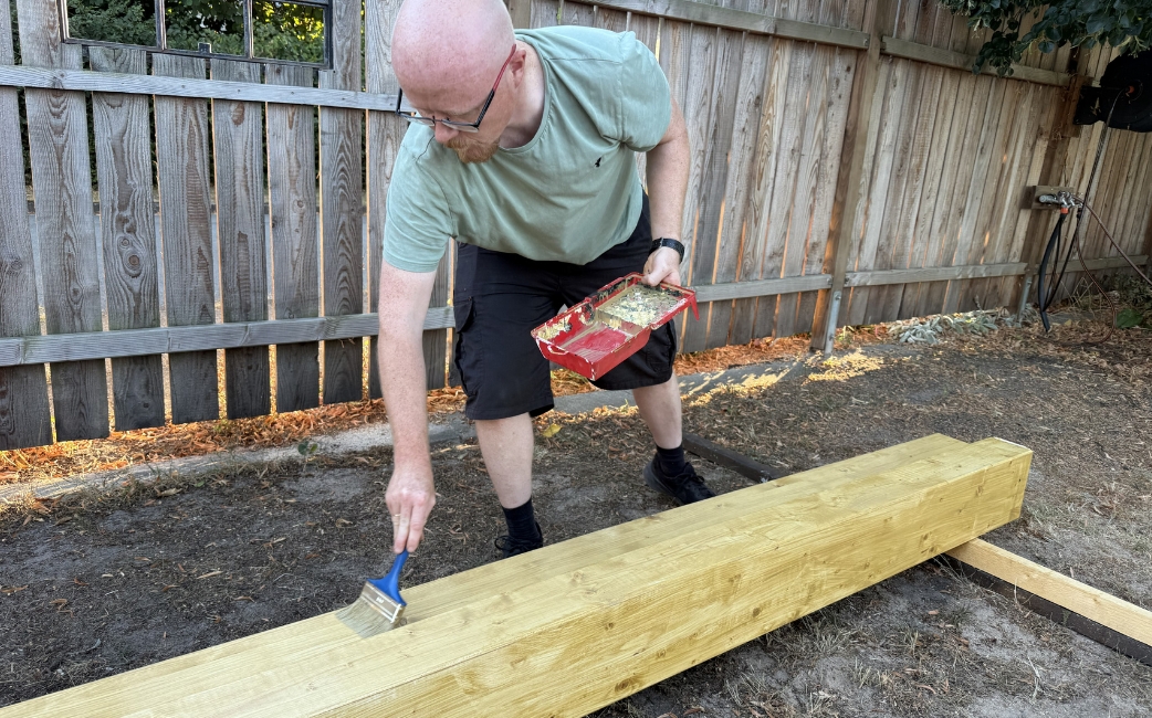 Eine Person trägt im Garten mit einem Pinsel Holzschutzmittel auf einen Holzbalken auf.