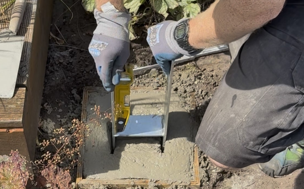 Eine Person, die Handschuhe trägt, bearbeitet mit einem gelben Handwerkzeug eine Metallhalterung, die in frischem Beton neben einer Holzkonstruktion verankert ist.