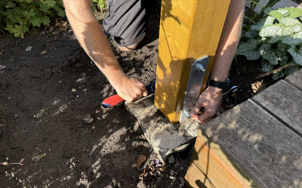 Eine Person befestigt einen Holzpfosten mithilfe einer Metallhalterung und eines Handwerkzeugs an einem Betonsockel.