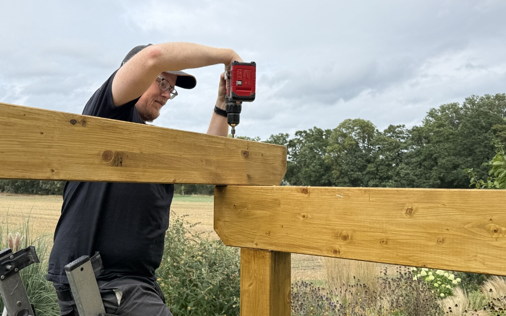  Eine Person befestigt mit einem Akku-Bohrer im Freien einen Holzbalken am Rahmen einer Pergola.