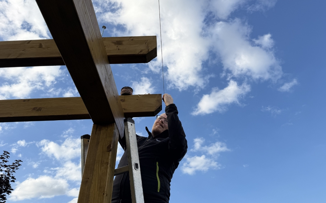 Eine Person auf einer Leiter montiert einen Holzbalken am oberen Teil eines Pergola-Rahmens.