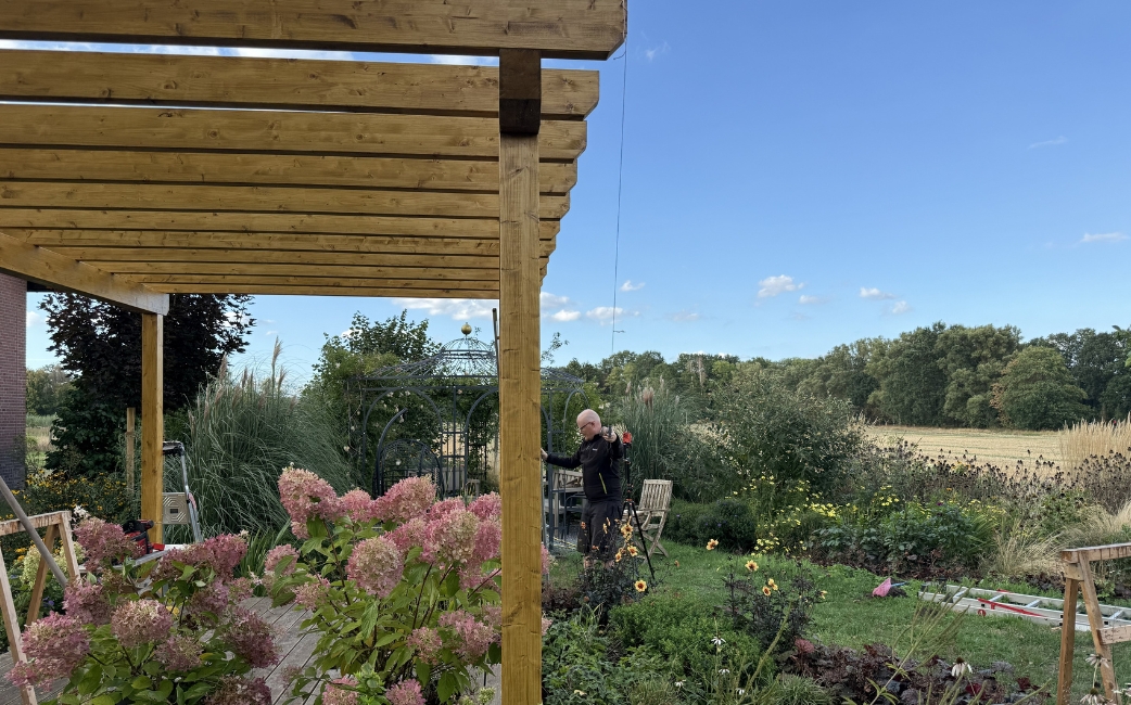 Fertiggestellte Holzpergola mit einem Lamellendach über einer Sitzecke im Garten, umgeben von blühenden Pflanzen und Grünflächen.