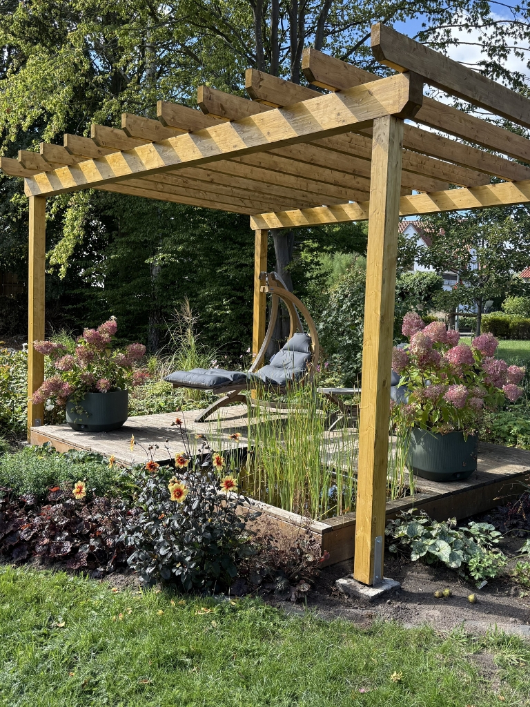 Eine Pergola aus Holz mit einer Sitzecke, Hortensien in Töpfen und blühenden Pflanzen in einem Garten.