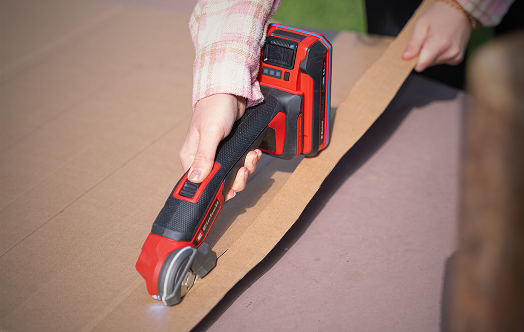 Hand hält roten Akku-Schneider und schneidet eine Kartonplatte auf einem Tisch.