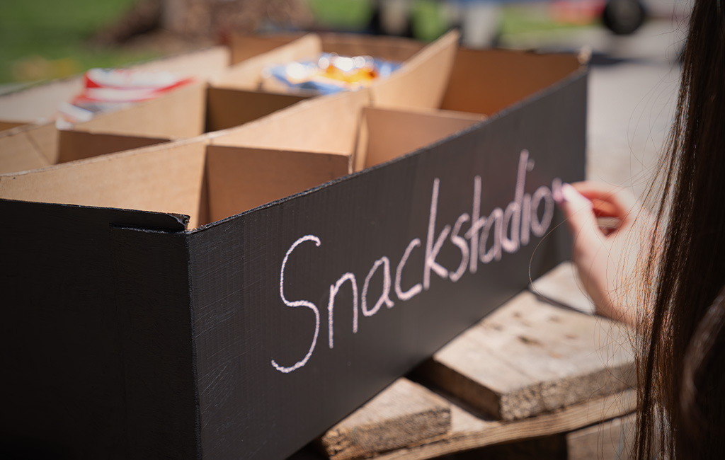 Hand schreibt mit weißer Kreide „Snackstadion“ auf eine schwarz gestrichene Box.