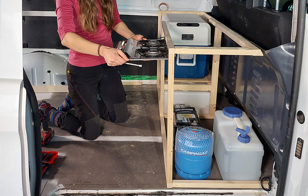 Eine Frau kniet im Transporter und hält ein Gaskochfeld, das in eine Holzkonstruktion eingebaut werden soll.