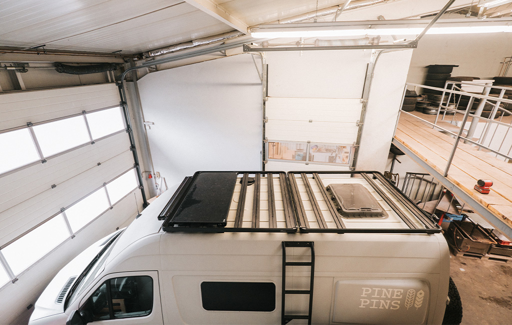 Camper van with an empty roof frame parked in a workshop under an open garage door.