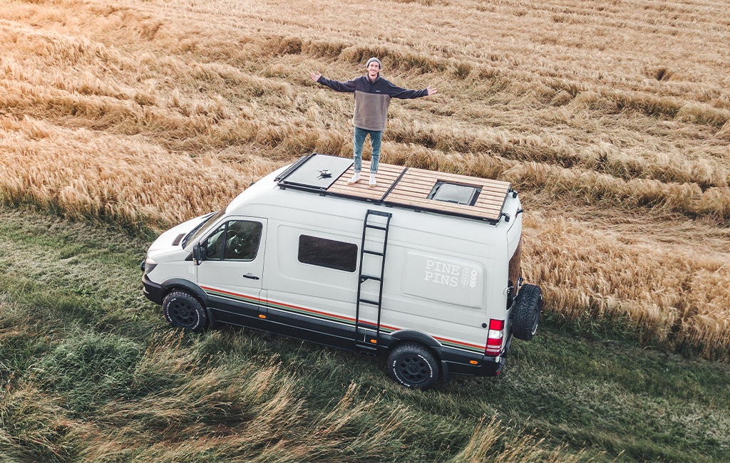 Person standing with arms outstretched on top of a camper van in the middle of a grain field.