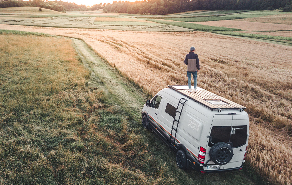 Camper van with a finished roof terrace driving along a field path at sunset.