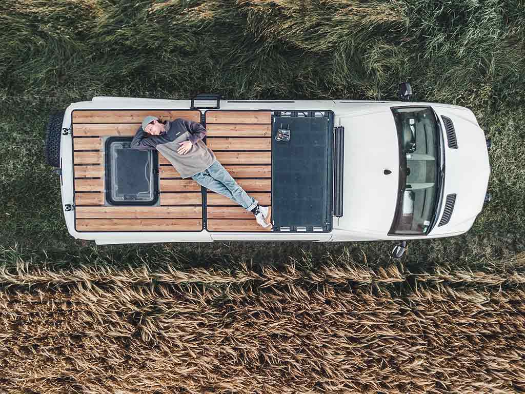 Person relaxing on a wooden rooftop terrace of a camper van next to a grain field.