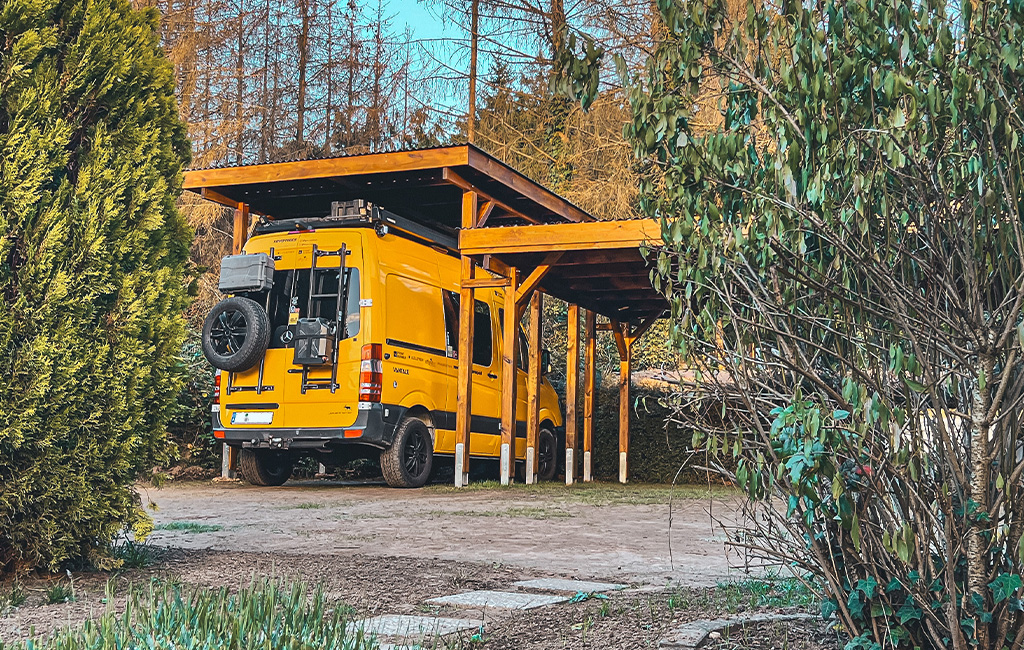 A yellow van parked under an open wooden carport, surrounded by trees and bushes.