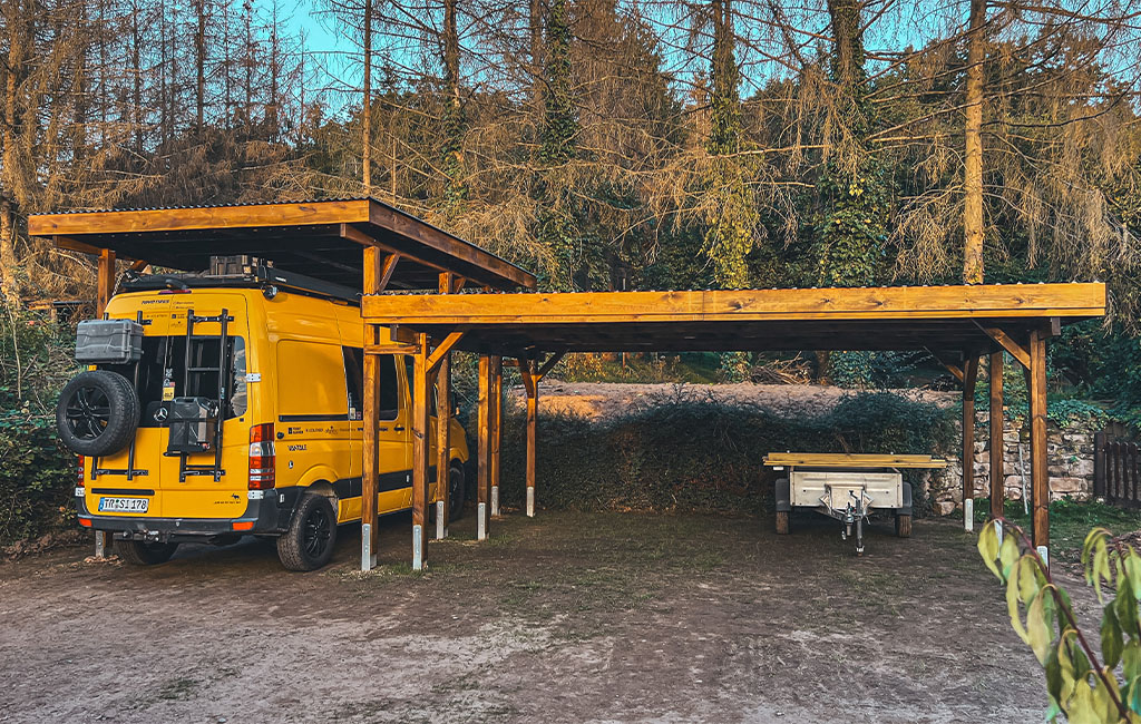 A yellow van and a trailer parked under a large, open wooden carport.