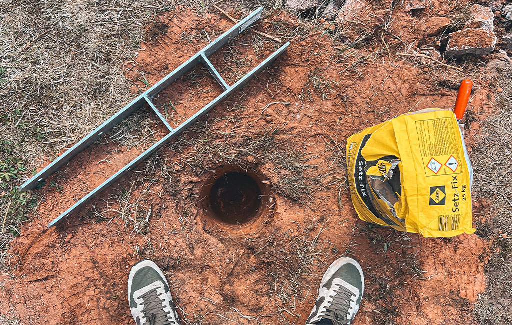A round hole in the ground, next to a metal beam, a yellow concrete bag, and visible shoes.