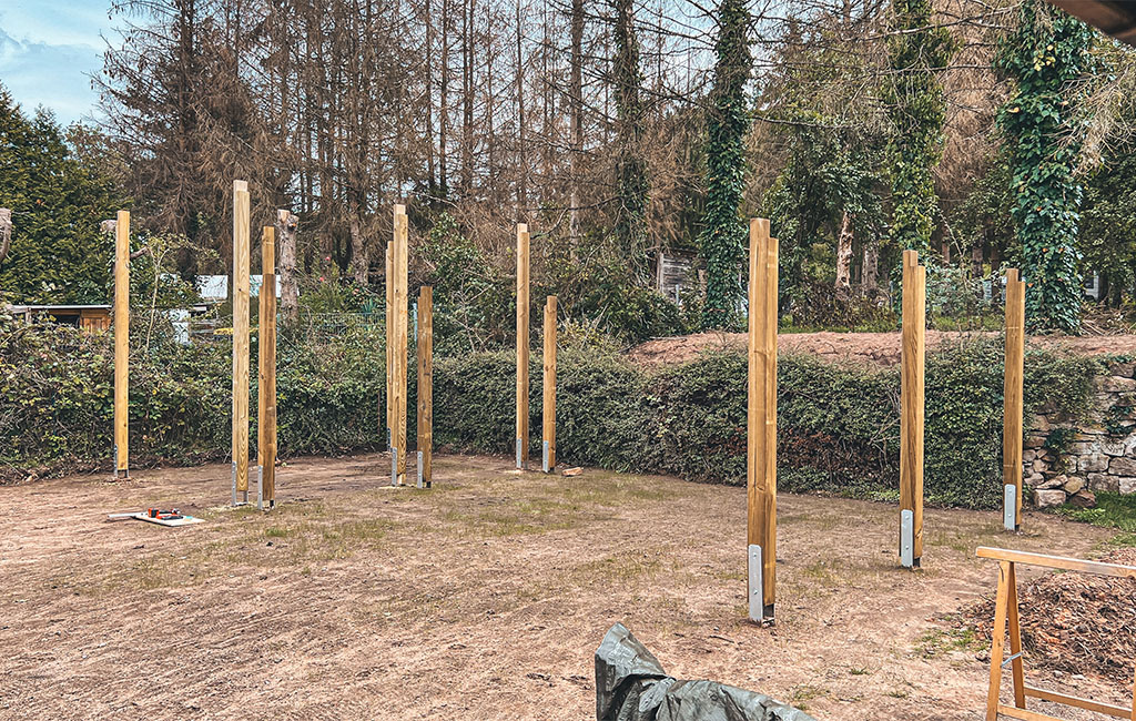 Several upright wooden posts with metal bases standing on a flat area, construction site in the background.