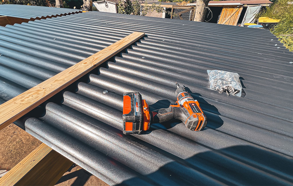 Corrugated roof of a carport with an Einhell cordless drill, wooden slat, and bag of screws on top.