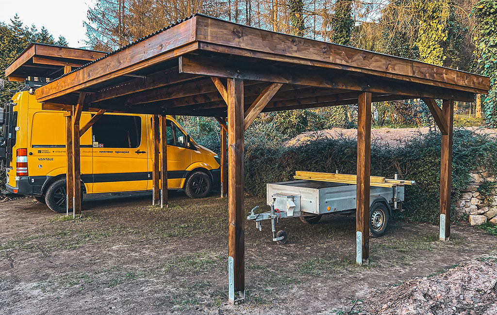 A yellow van and a trailer parked under a finished, brown-painted wooden carport.