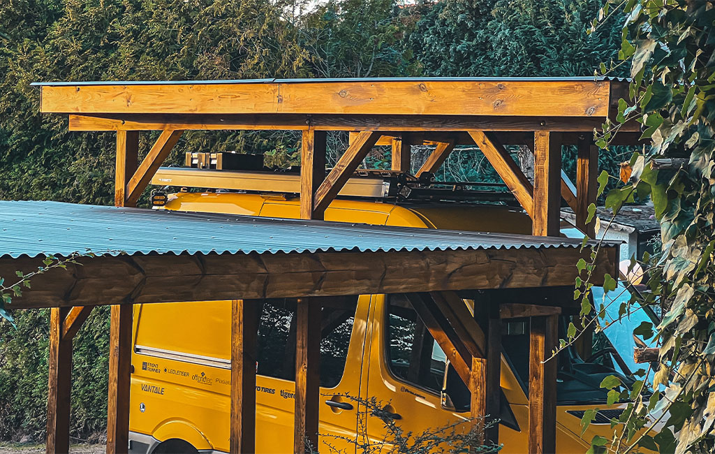 Close-up of the wooden structure of the carport, a yellow van in the background.