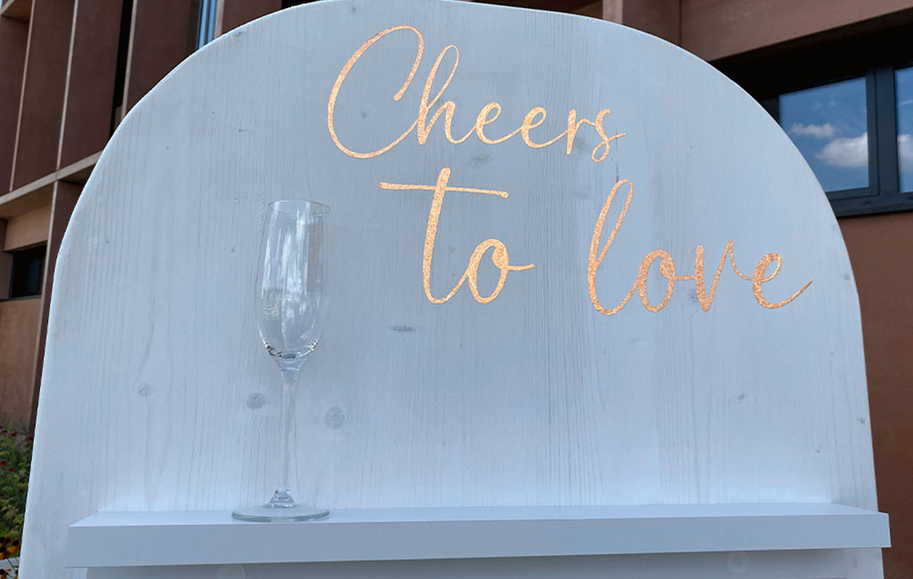 Nahaufnahme des oberen Bereichs einer weiß gestrichenen DIY-Sektwand mit abgerundetem Abschluss, goldfarbener Aufschrift „Cheers to love“ und einem Sektglas auf der Ablage.