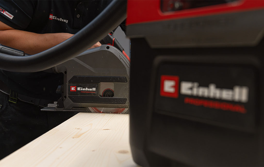 Eine Person schneidet mit einer Einhell Professional Handkreissäge eine Holzplatte zu; ein angeschlossener Absaugschlauch reduziert den Staub.