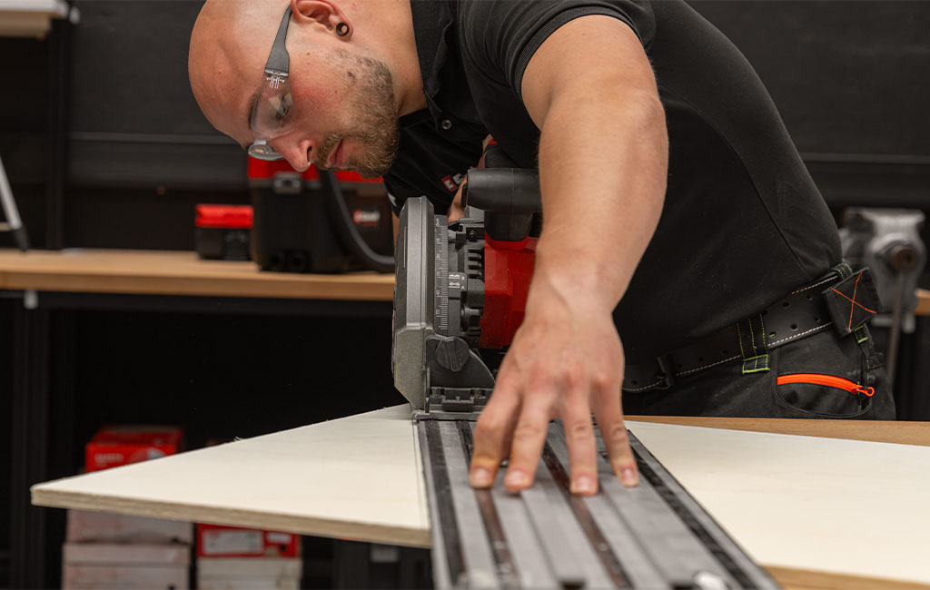 Ein Handwerker mit Schutzbrille schneidet eine Holzplatte mit einer Einhell Tauchsäge entlang einer Führungsschiene zu.