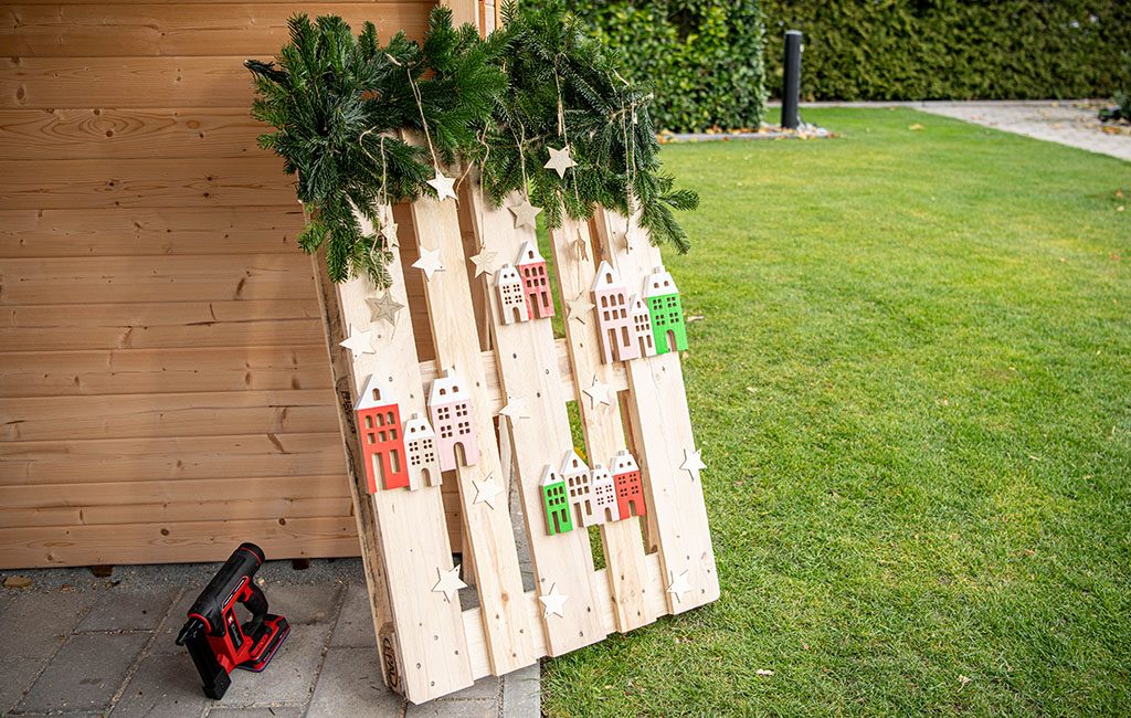 A Christmas-themed pallet with wooden houses, stars, and fir branches leans against a wooden shed, next to an Einhell stapler.