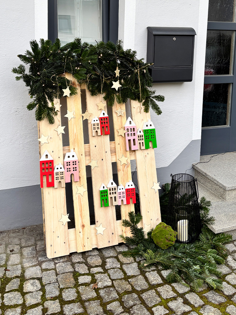 Decorative wooden pallet with stars, wooden houses, and fir branches stands in front of a house entrance on cobblestones.
