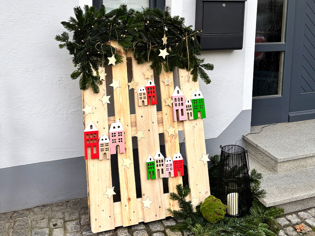 Decorative wooden pallet with stars, wooden houses, and fir branches stands in front of a house entrance on cobblestones.