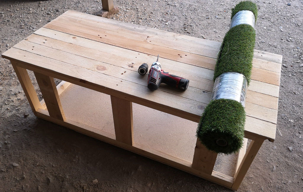 A simple wooden table stands in a workshop. On the table are a drill and a roll of artificial grass.