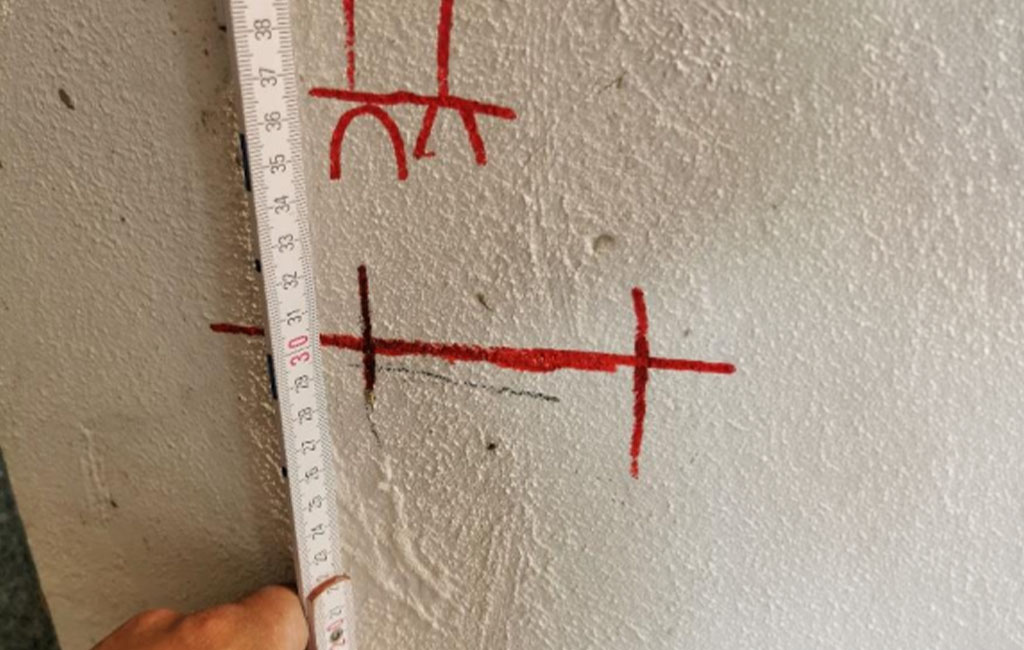 Hand holds a measuring tool against a wall with red markings for drilling points and distances.