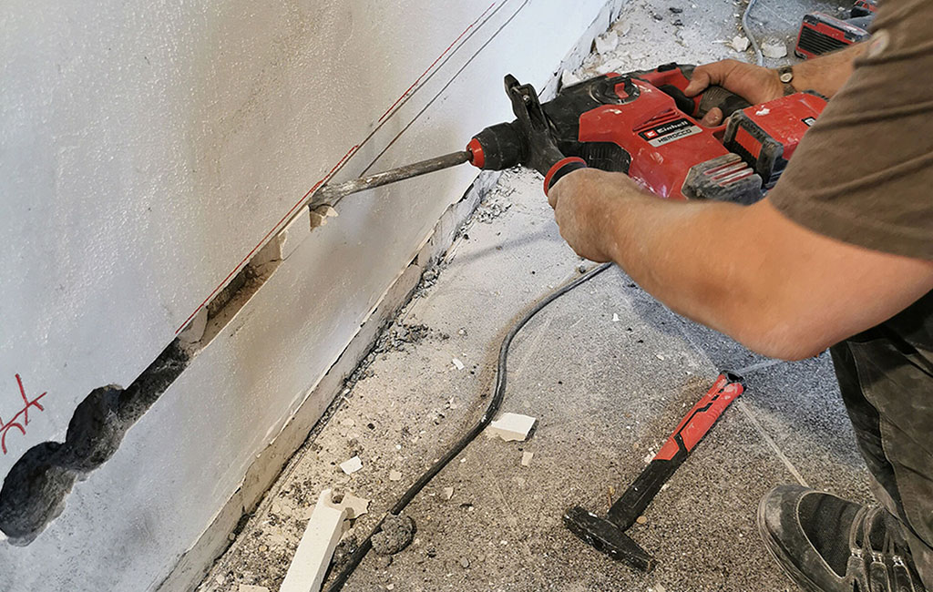 Hand guides a drill along a guide in a wall groove, with dust and debris below.