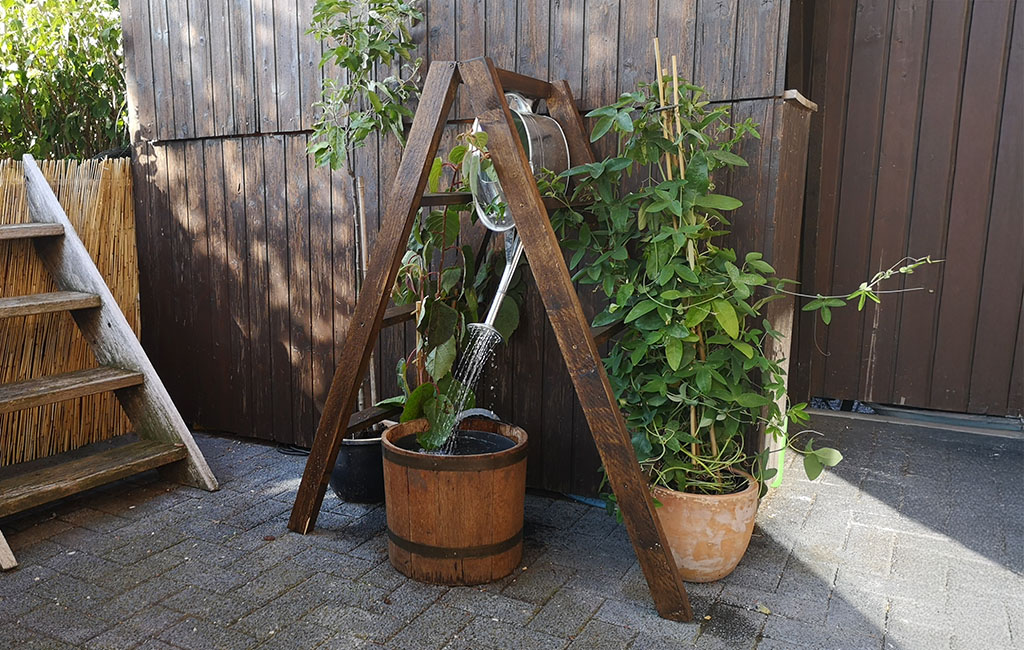 A self-made water feature with a wooden frame, watering can, and plants on a patio.