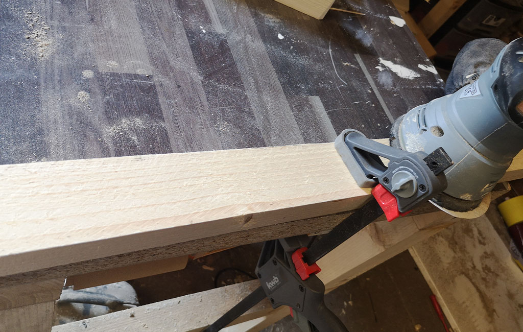 A sanding machine smooths a wooden board, fixed to the table with a clamp.
