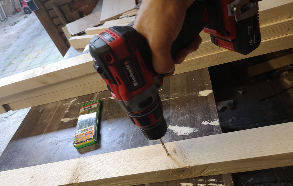 A hand drills a hole into a wooden slat using an Einhell cordless drill, a bit set is lying next to it.